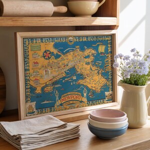May include: A framed vintage map of Newport, Rhode Island, in a wooden frame. The map is in shades of blue, yellow, and brown. A stack of beige linen napkins, a small stack of pastel bowls, and a vase of flowers are also visible.