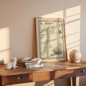 May include: A framed vintage map of Syria, with detailed cartography in beige and brown tones. The map is displayed on a wooden desk, accompanied by a white ceramic cat figurine, books, and a decorative vase.