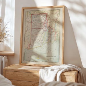 Pode incluir: Um mapa vintage emoldurado de Rhode Island, com cartografia detalhada em tons suaves de bege, verde e vermelho. O mapa está emoldurado em uma moldura de madeira clara, exibido em uma cômoda de madeira. O título do mapa, "Rhode Island", é visível.