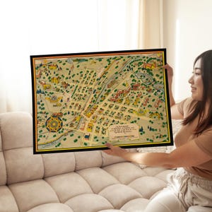 Puede incluir: Una impresión de mapa vintage enmarcada. El mapa presenta una ilustración detallada de una ciudad con edificios, árboles y un río. La impresión tiene un borde amarillo y texto en la parte inferior.