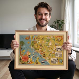 May include: A framed vintage-style illustrated map titled Cote de Beaute, featuring a yellow background with detailed drawings of coastal landmarks, ships, and figures, enclosed in a light-colored wooden frame with a cream-colored mat border.