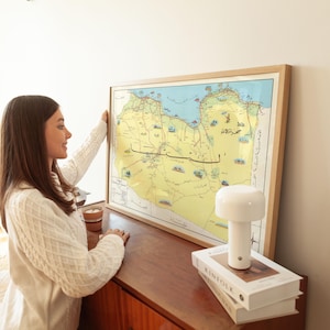 May include: A framed map of a region with yellow, blue, and brown tones. The map is in a light wood frame. A white lamp and two books are on a wooden surface. A person is holding the map.