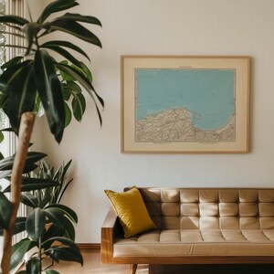 Può includere: Una mappa vintage di una regione costiera è incorniciata e appesa a un muro bianco sopra un divano in pelle marrone con un cuscino giallo.