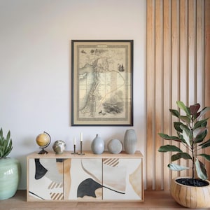 May include: A framed antique map hangs above a wooden cabinet with abstract art on the doors. The cabinet is adorned with globes, candles, and vases. A potted plant is on the right. The map shows a region with a red line.