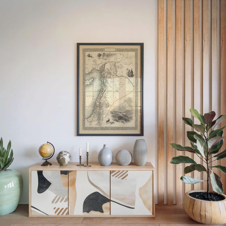 May include: A framed antique map hangs above a wooden cabinet with abstract art on the doors. The cabinet is adorned with globes, candles, and vases. A potted plant is on the right. The map shows a region with a red line.
