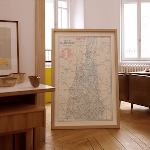May include: A framed vintage map of New Hampshire, featuring a detailed road network and geographical features. The map is in a light-colored frame, displayed on a wooden floor. The text "New Hampshire" is visible at the top of the map.