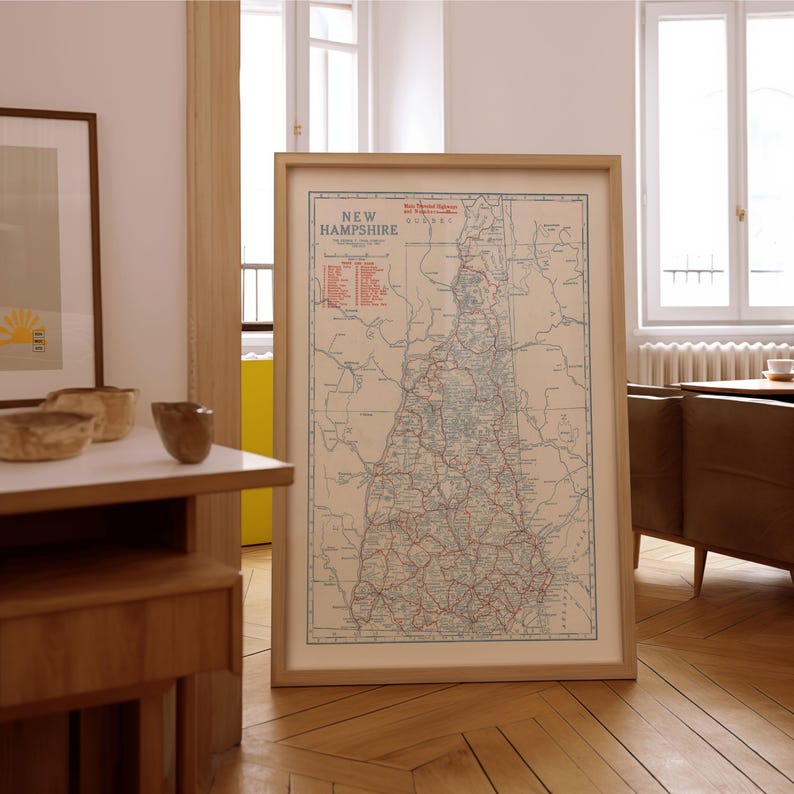 May include: A framed vintage map of New Hampshire, featuring a detailed road network and geographical features. The map is in a light-colored frame, displayed on a wooden floor. The text "New Hampshire" is visible at the top of the map.