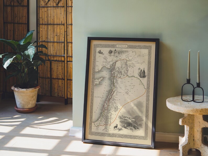 May include: A framed antique map of Syria, displaying detailed geographical features and historical illustrations. The map is in a black frame, leaning against a pale green wall. A potted plant and a bamboo screen are in the background.