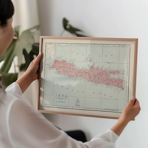 Peut inclure: Carte ancienne encadrée de Java, Indonésie, dans un cadre en bois clair. La carte présente l'île avec des terres rouges et roses sur un fond bleu pâle. Le texte "JAVA" est visible en bas.