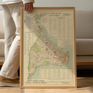 May include: A framed vintage map of Constantinople, featuring detailed city streets, green park areas, and blue water bodies. The map has text in French and is encased in a light brown wooden frame. The map is held by a person.