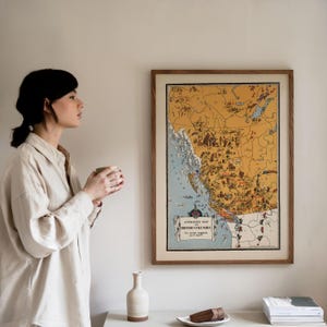 May include: A framed wall art print featuring an illustrated map titled Animated Map of British Columbia. The map uses a mustard yellow background for land and light blue for water, filled with small drawings of animals and landmarks.