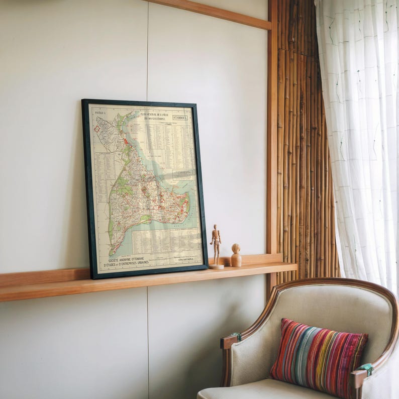 May include: Framed vintage map with a black frame. The map features detailed cartography with red, green, and beige tones. The map is displayed on a wooden shelf with small wooden figurines. A colorful striped pillow rests on a chair.