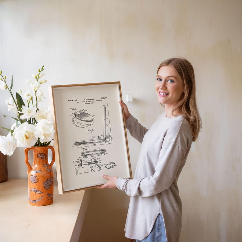 May include: A framed patent print featuring technical drawings in black and white. The print is held by a person. A decorative orange vase with white flowers is on a wooden surface.