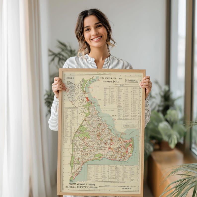 May include: A framed vintage map of Istanbul, Turkey, with detailed city plans and text in French. The map features a beige background with green, red, and blue accents, encased in a light brown wooden frame. The map is held up by a person.
