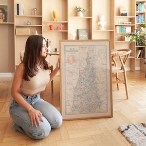 May include: A framed vintage map of New Hampshire with a beige border. The map features red and blue lines representing roads and waterways. The title "New Hampshire" is printed at the top. The map is displayed in a light-colored wooden frame.