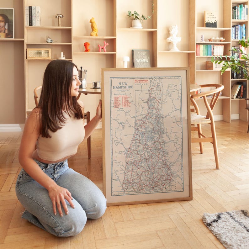 May include: A framed vintage map of New Hampshire with a beige border. The map features red and blue lines representing roads and waterways. The title "New Hampshire" is printed at the top. The map is displayed in a light-colored wooden frame.