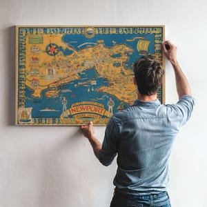 May include: A framed vintage map of Newport, Rhode Island, in a rectangular wooden frame. The map features a detailed illustration of the city with a nautical theme, including ships and a compass rose. The map is held by a person.