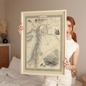May include: A framed vintage map of Syria, displaying detailed cartography with ornate borders and illustrations. The map is presented in a light-coloured frame, highlighting the historical geography of the region. The word "SYRIA" is at the top of the map.
