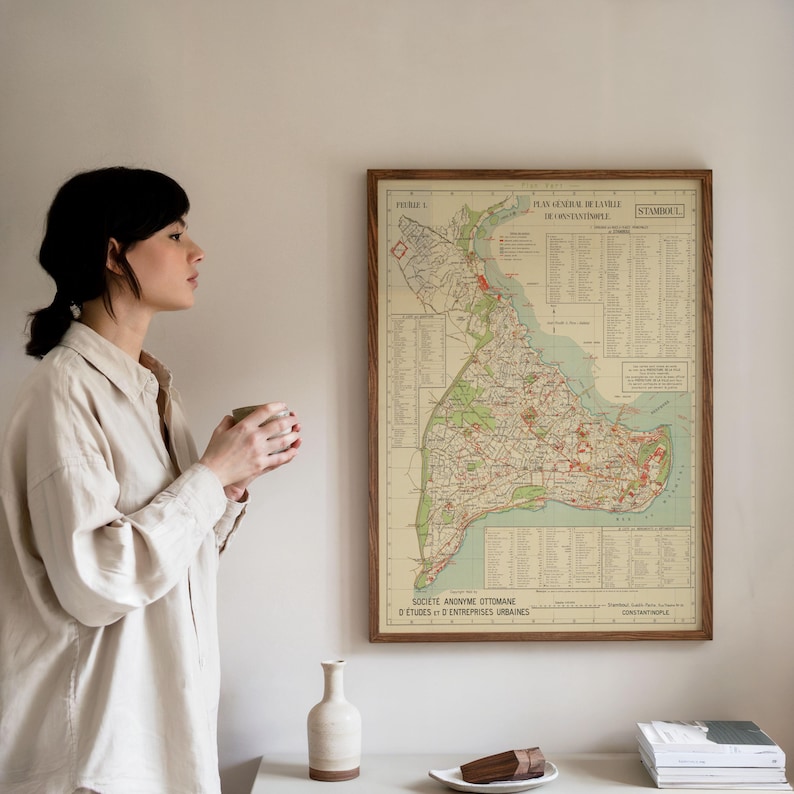 May include: A framed vintage map of Istanbul, Turkey, with a wooden frame. The map features detailed city streets and landmarks in shades of green, red, and beige. The map is hanging on a white wall.