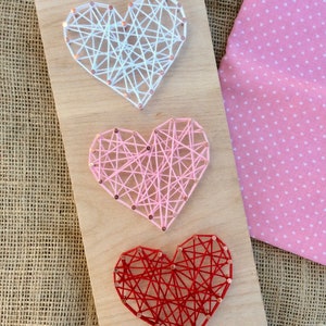 Könnte beinhalten: Drei String Art Herzen auf einem Holzbrett. Das obere Herz ist weiß, das mittlere Herz ist rosa und das untere Herz ist rot.