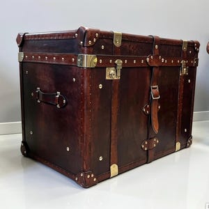 Handmade dark brown leather steamer trunk with brass corner plates, stud trim and leather carry straps, photographed between two leather armchairs