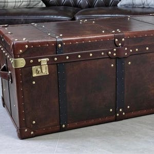 Puede incluir: Un baúl rectangular de cuero marrón oscuro con correas de cuero negro, refuerzos de metal en las esquinas en tono latón y dos cierres en tono latón en la parte frontal. El baúl presenta pequeños remaches en tono latón.
