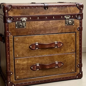 May include: A brown leather-look storage chest featuring two drawers with dark brown leather strap handles. The exterior is accented with dark brown leather trim, brass-toned metal corner reinforcements, decorative studs, and two brass-toned metal locks.