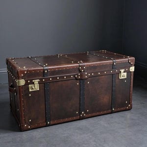 Puede incluir: Un baúl de almacenamiento rectangular de color marrón oscuro con correas de cuero negro, refuerzos de esquina de metal en tono latón y dos pestillos de latón en la parte frontal. El exterior presenta numerosos remaches de latón pequeños.