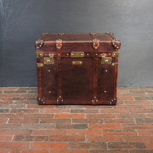 Op de afbeelding: Een vintage bruin leren koffer met messing hardware, waaronder hoekbeschermers, grendels en decoratieve platen. De koffer is rechthoekig en staat op een bakstenen vloer.