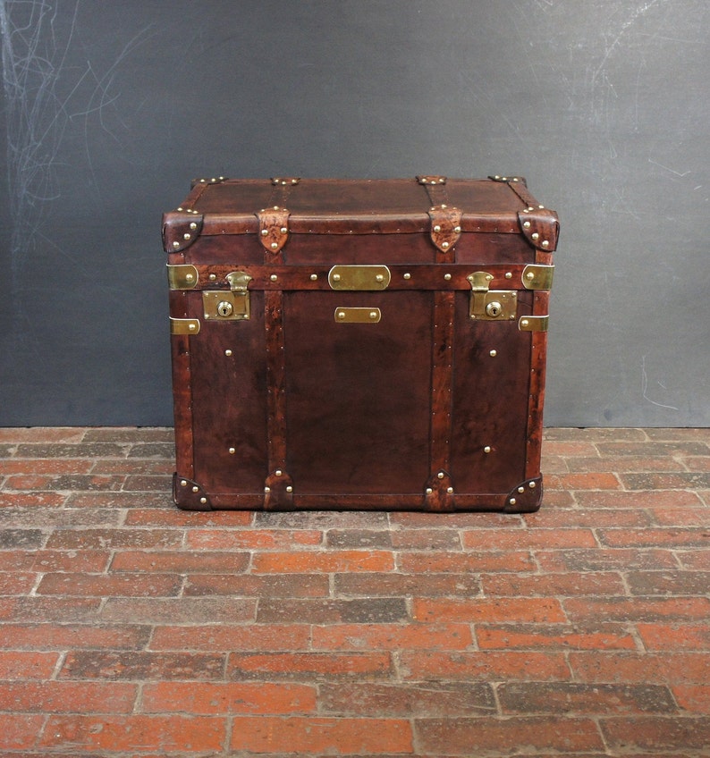Op de afbeelding: Een vintage bruin leren koffer met messing hardware, waaronder hoekbeschermers, grendels en decoratieve platen. De koffer is rechthoekig en staat op een bakstenen vloer.