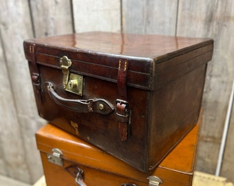May include: A vintage brown leather suitcase with a brass lock and leather straps. The suitcase is sitting on top of a smaller brown suitcase.