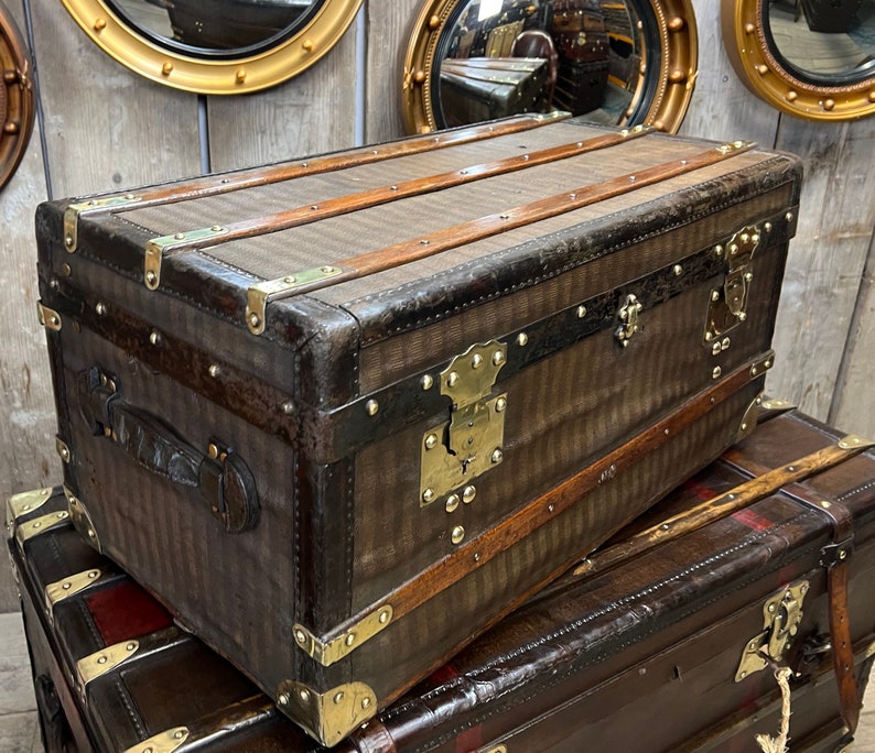 Puede incluir: Un ba&uacute;l marr&oacute;n vintage con herrajes de lat&oacute;n y detalles de cuero. El ba&uacute;l presenta un patr&oacute;n de rayas en la parte delantera y superior, con protectores de esquina de lat&oacute;n y una cerradura central. El ba&uacute;l es rectangular.