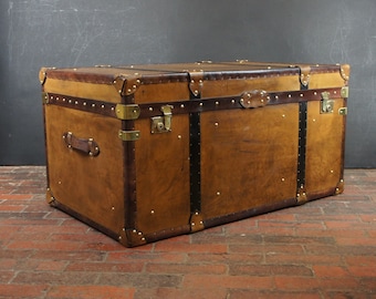 May include: A large rectangular storage trunk featuring a tan leather-like exterior with dark brown leather straps, brass-toned metal corner reinforcements, and brass-toned latches. The trunk includes a side handle and decorative metal studs.