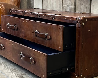 Könnte beinhalten: Eine mit braunem Leder bezogene Kommode mit zwei ausziehbaren Schubladen und dunklen Ledergriffen. Das Möbelstück ist an den Kanten und Ecken mit messingfarbenen Metallnieten verziert, und die Schubladenfronten sind mit passenden Metallbeschlägen befestigt.