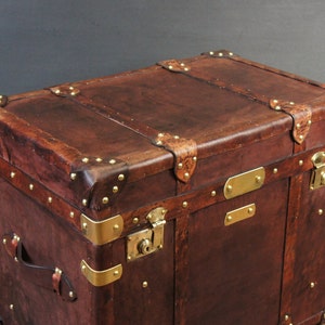 Op de afbeelding: Een bruine leren koffer met messing details. De koffer heeft messing hoekbeschermers, sloten en decoratieve banden. Het leer vertoont tekenen van slijtage, wat het een vintage uitstraling geeft. De koffer is rechthoekig en robuust.