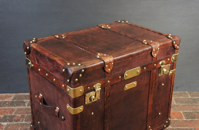 Op de afbeelding: Een bruine leren koffer met messing details. De koffer heeft messing hoekbeschermers, sloten en decoratieve banden. Het leer vertoont tekenen van slijtage, wat het een vintage uitstraling geeft. De koffer is rechthoekig en robuust.