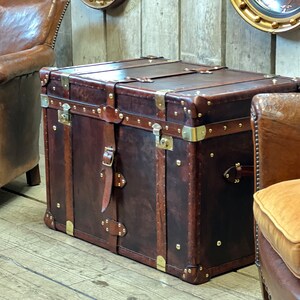 Dark brown leather steamer chest with solid brass fittings and stud trim, styled as a coffee table between two studded leather chairs