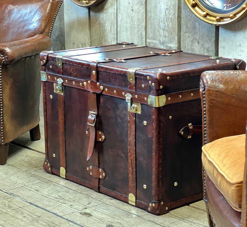 Dark brown leather steamer chest with solid brass fittings and stud trim, styled as a coffee table between two studded leather chairs