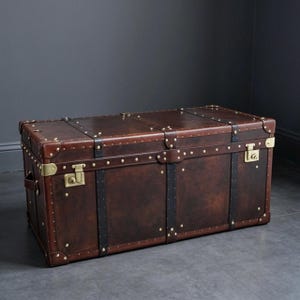 Peut inclure: Un coffre de rangement rectangulaire en cuir marron foncé, doté de sangles en cuir noir, de renforts d'angle en métal doré et de clous décoratifs en laiton. Le coffre comprend deux loquets en laiton sur le devant et des poignées latérales.