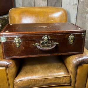 Peut inclure: Une valise rectangulaire marron avec une finition texturée façon crocodile. La valise présente des ferrures en laiton poli, incluant deux loquets latéraux, un mécanisme de verrouillage central, des coins renforcés et une poignée en cuir.