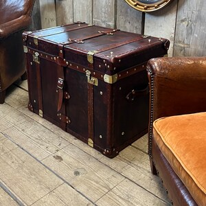 Handcrafted English leather trunk with brass corner guards, leather carry straps and central locking clasp on a white floor
