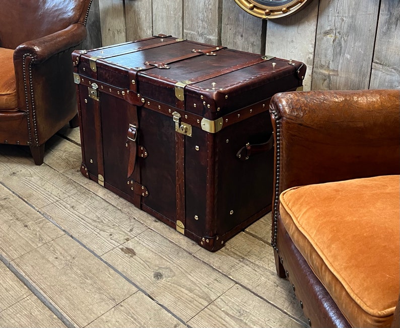 Handcrafted English leather trunk with brass corner guards, leather carry straps and central locking clasp on a white floor