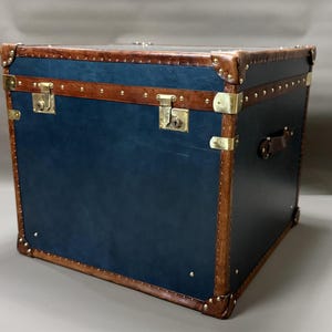 May include: A square storage trunk featuring a deep blue exterior with brown leather trim along the edges. The trunk is accented with polished brass corner protectors, metal studs, and two front-facing locking latches with keyholes.