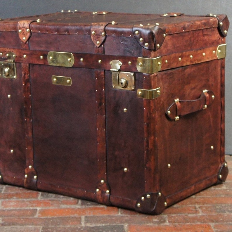 Op de afbeelding: Een grote, antieke bruin lederen koffer met messing hardware. De koffer heeft versterkte hoeken, messing studs en een slot. Een leren handvat is zichtbaar aan de zijkant. De koffer is een vintage opbergoplossing.