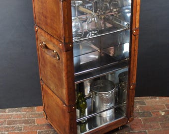 Puede incluir: Un mueble bar de cuero marrón de estilo vintage con detalles de latón. El mueble tiene una puerta de cristal que revela estantes con cristalería, una botella de vino y una cubitera de metal. Tiene un asa y ruedas. Mide unos 100 cm de alto.
