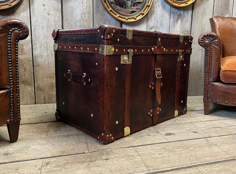 Dark hand dyed leather trunk with brass stud trim, corner plates and buckled carry straps, used as a coffee table chest