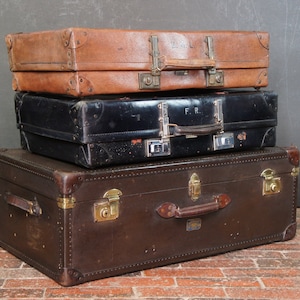 Może przedstawiać: Trzy vintage'owe walizki ułożone jedna na drugiej. Górna walizka jest ze skóry brązowej z mosiężnymi okuciami. Środkowa walizka jest ze skóry czarnej z mosiężnymi okuciami i inicjałami "F.R." z przodu. Dolna walizka jest ze skóry brązowej z mosiężnymi okuciami i skórzanym uchwytem.