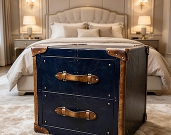 Puede incluir: Una mesita de noche azul marino con dos cajones, asas y esquinas de cuero marrón. La mesa tiene una superficie texturizada y está colocada sobre una alfombra blanca y esponjosa. El diseño evoca una estética de maleta de viaje vintage.