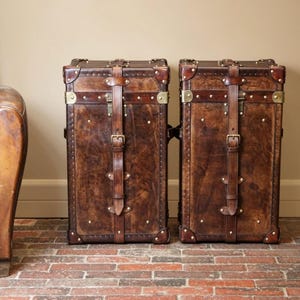 Puede incluir: Dos baúles de cuero marrón de estilo vintage con herrajes de latón y correas de cuero. Los baúles son rectangulares y verticales, con un aspecto envejecido. Están colocados sobre un suelo de ladrillo, sugiriendo un tema de viaje o almacenamiento clásico.