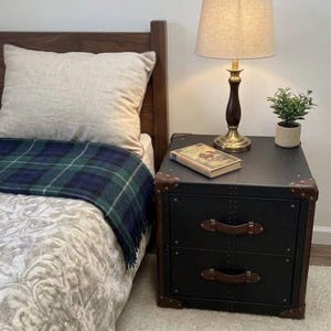Puede incluir: Una mesita de noche negra con dos cajones, asas de cuero marrón y detalles en las esquinas. Una lámpara con base de latón y pantalla beige, una pequeña planta y un libro encima. Una cama con una manta a cuadros y una almohada en el fondo.